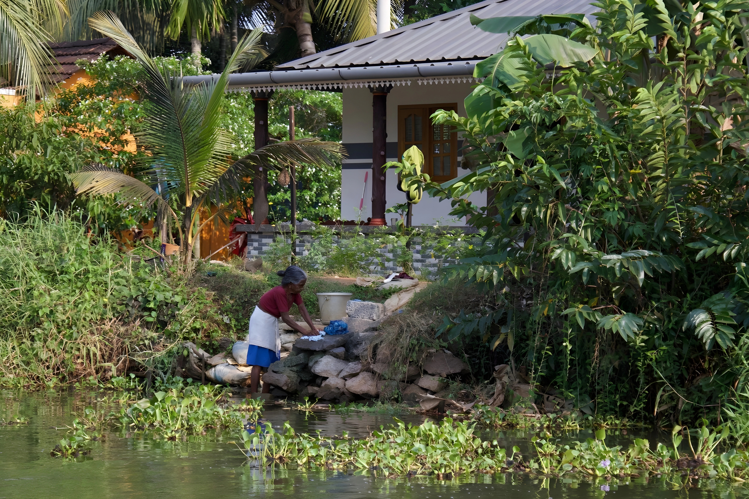 Woman in traditional attire washing clothes by the water's edge near a house surrounded by lush greenery, representing community resilience and self-sustainability.