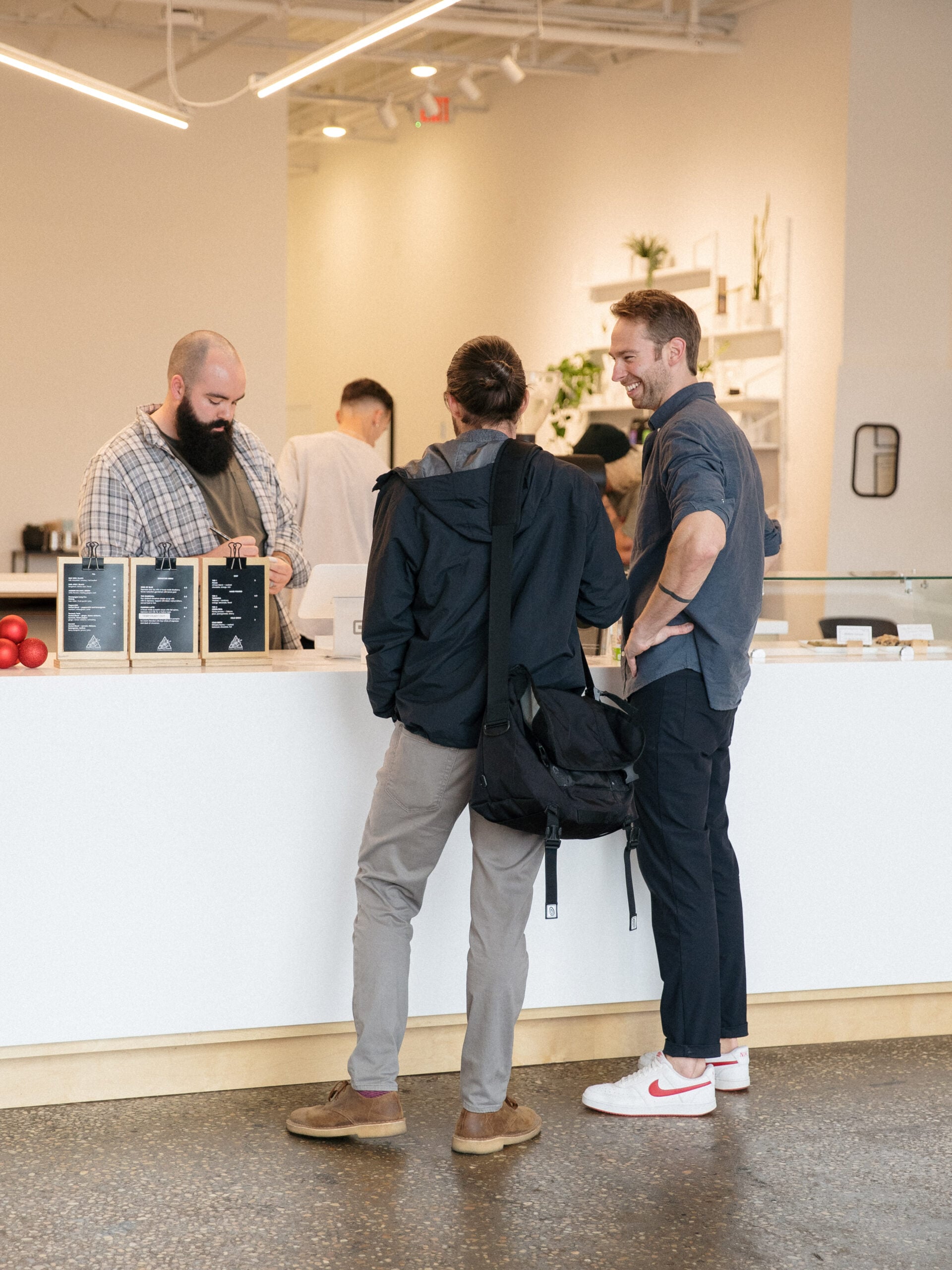 Two men engaged in conversation at a minimalist coffee shop counter, one with a beard wearing a plaid shirt, the other smiling in a casual outfit, while a barista prepares drinks in the background.