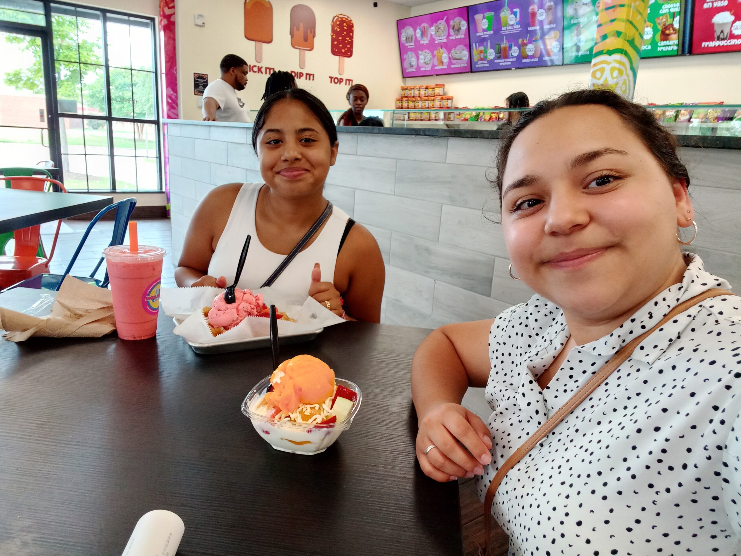Two young women enjoying ice cream and a drink at a dessert shop, with colorful decor and menu items visible in the background, highlighting community and celebration.