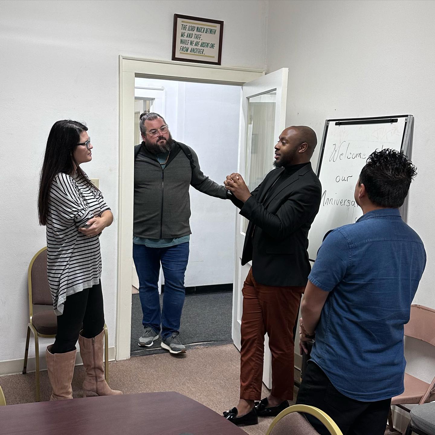 Group discussion in a meeting room, featuring a speaker in a black blazer, engaging with three attendees, including a woman in a striped shirt and tall boots, a man with a beard, and another man in a blue shirt, with a whiteboard displaying "Welcome to our Anniversary" in the background.
