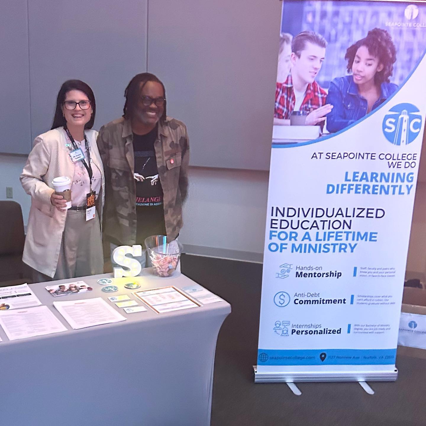 Two individuals smiling at a table with informational materials about Seapointe College, featuring a banner promoting individualized education for ministry, emphasizing mentorship and anti-debt commitment.