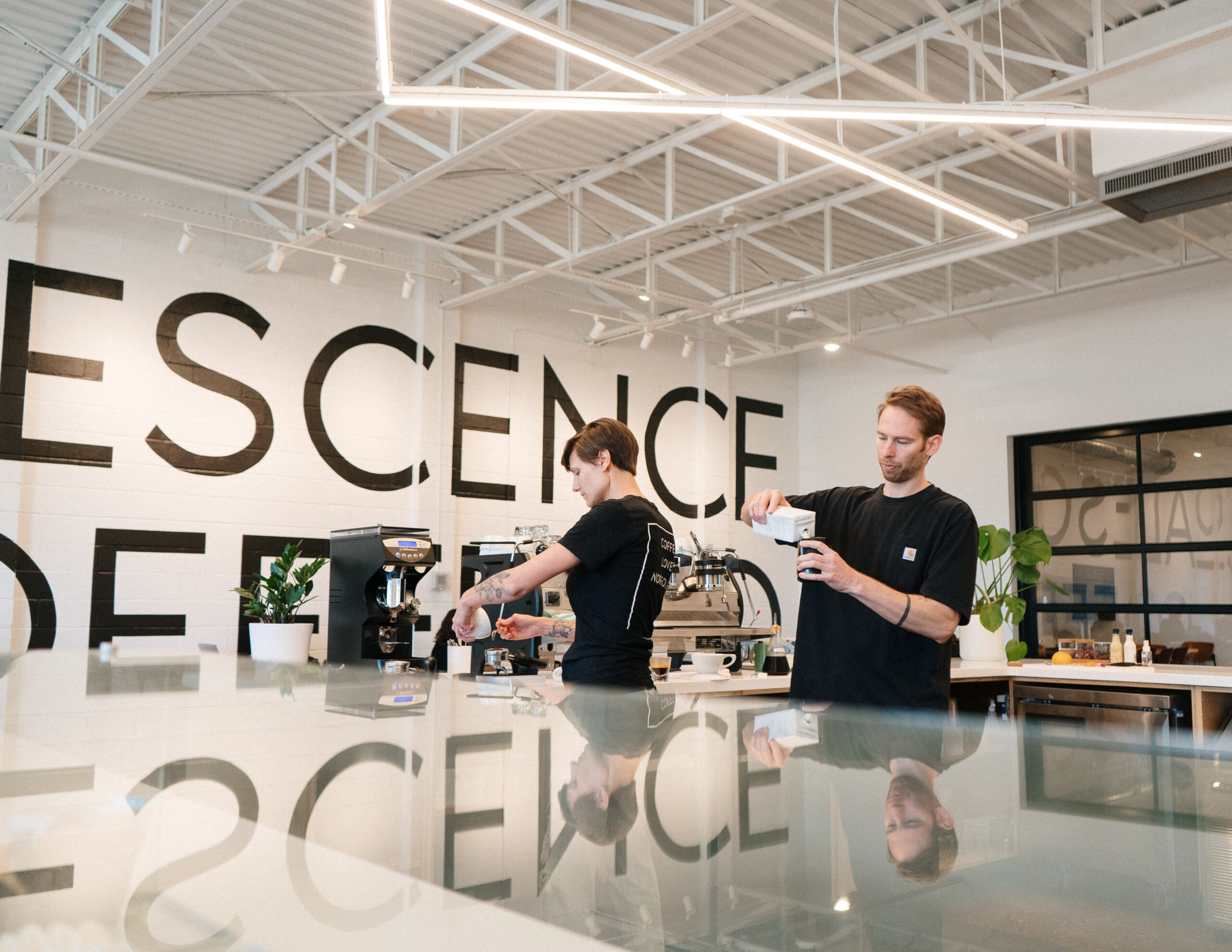 Baristas preparing coffee in a modern café with a large wall displaying the phrase "ESSENCE DEEP," emphasizing community and connection in a space focused on nurturing relationships and spiritual growth.