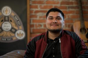 Young man smiling in a maroon jacket, seated in a cozy setting with exposed brick walls and a guitar in the background, representing community leadership in church planting efforts for Iglesia Mision Global.