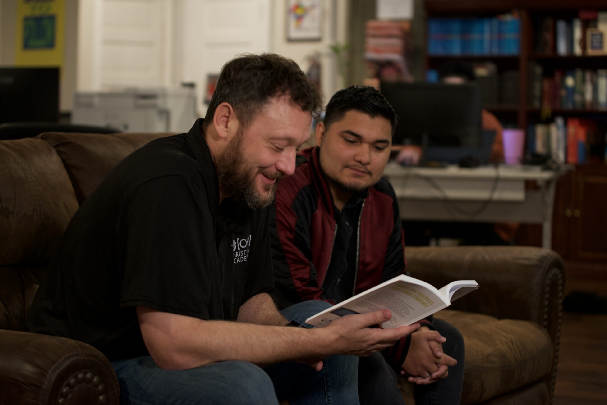 Two men engaged in discussion, one reading a book, in a cozy setting, symbolizing mentorship and community support in church planting efforts.