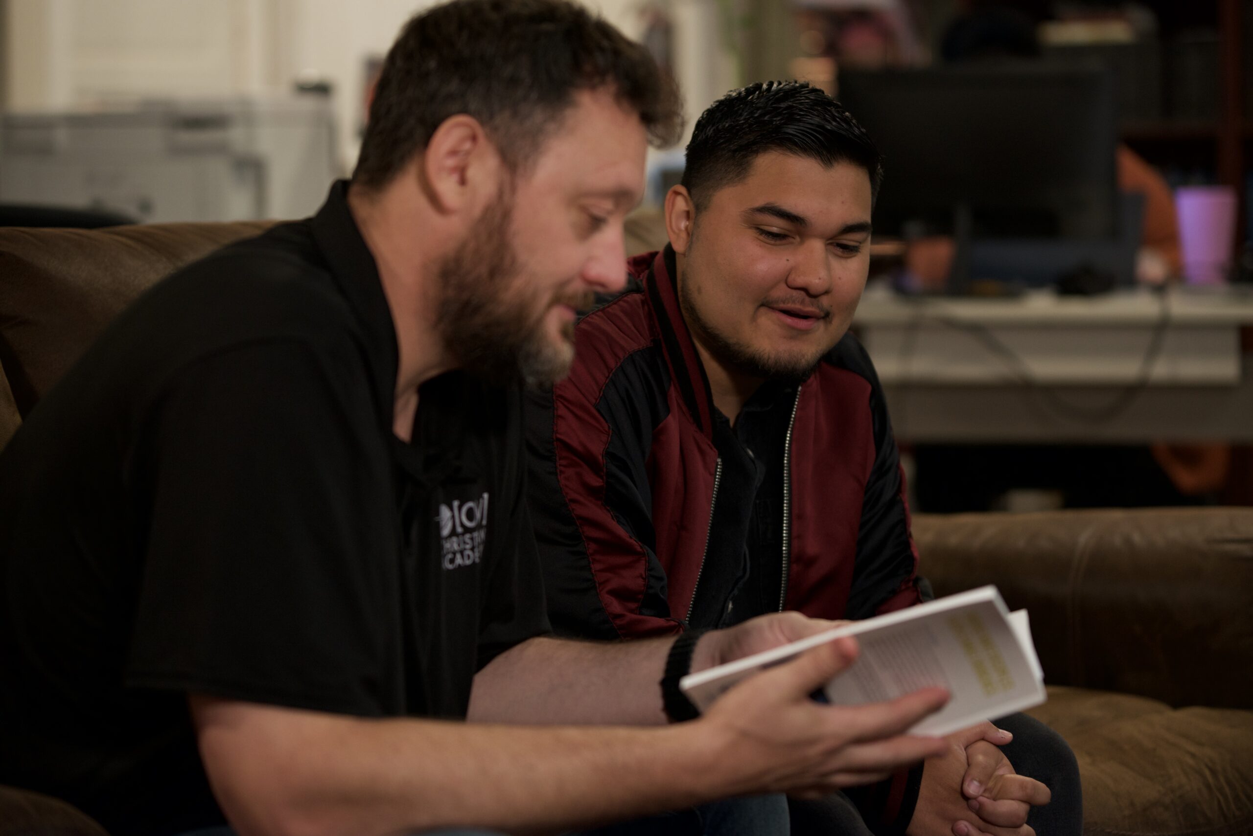 Two men engaged in discussion, one reading from a book, seated on a couch, reflecting the supportive environment of church planting and mentorship in Hispanic ministry.