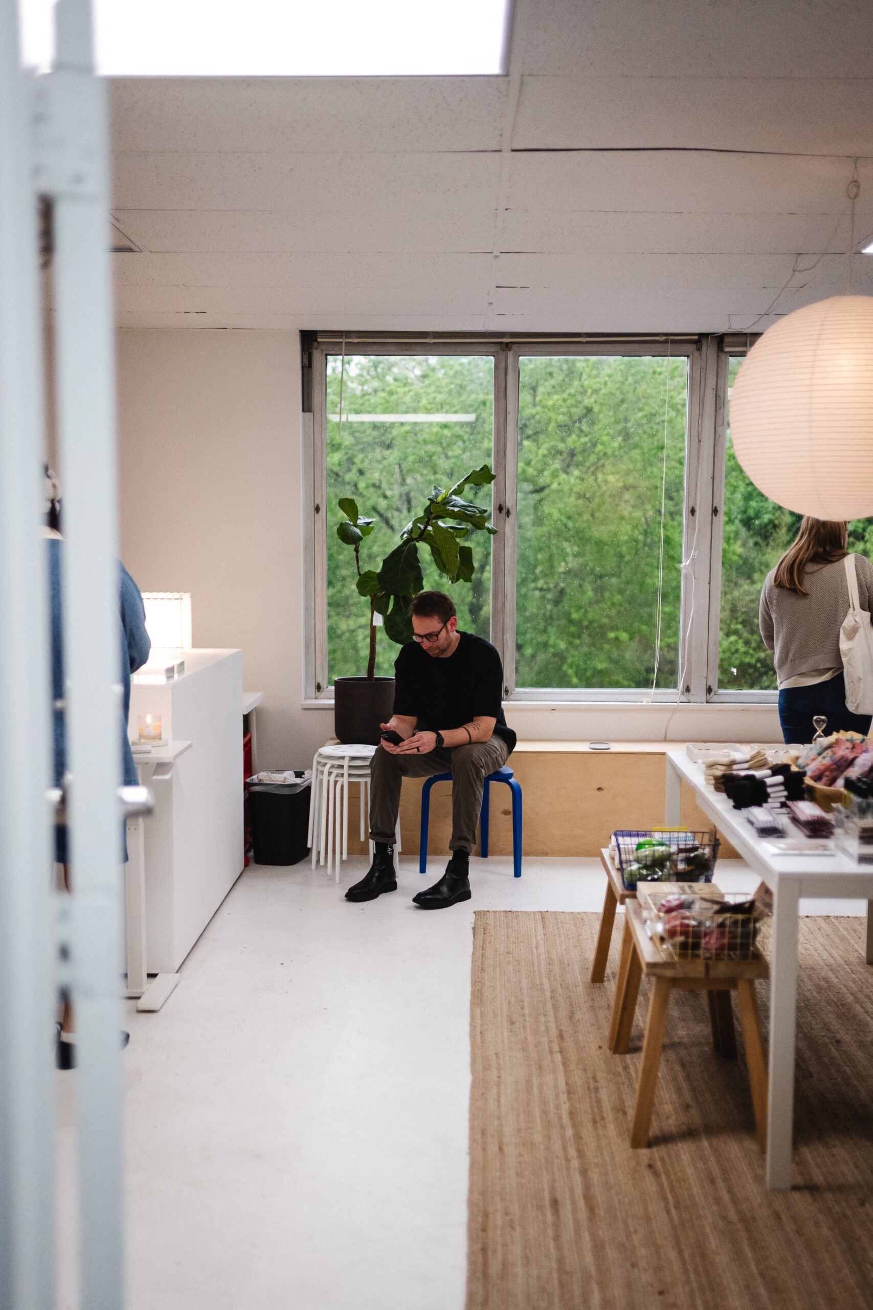 Man sitting on a blue chair in a bright, minimalist space, using a smartphone, with a potted plant and tables displaying various items, reflecting a community-focused environment for discipleship and church planting initiatives.