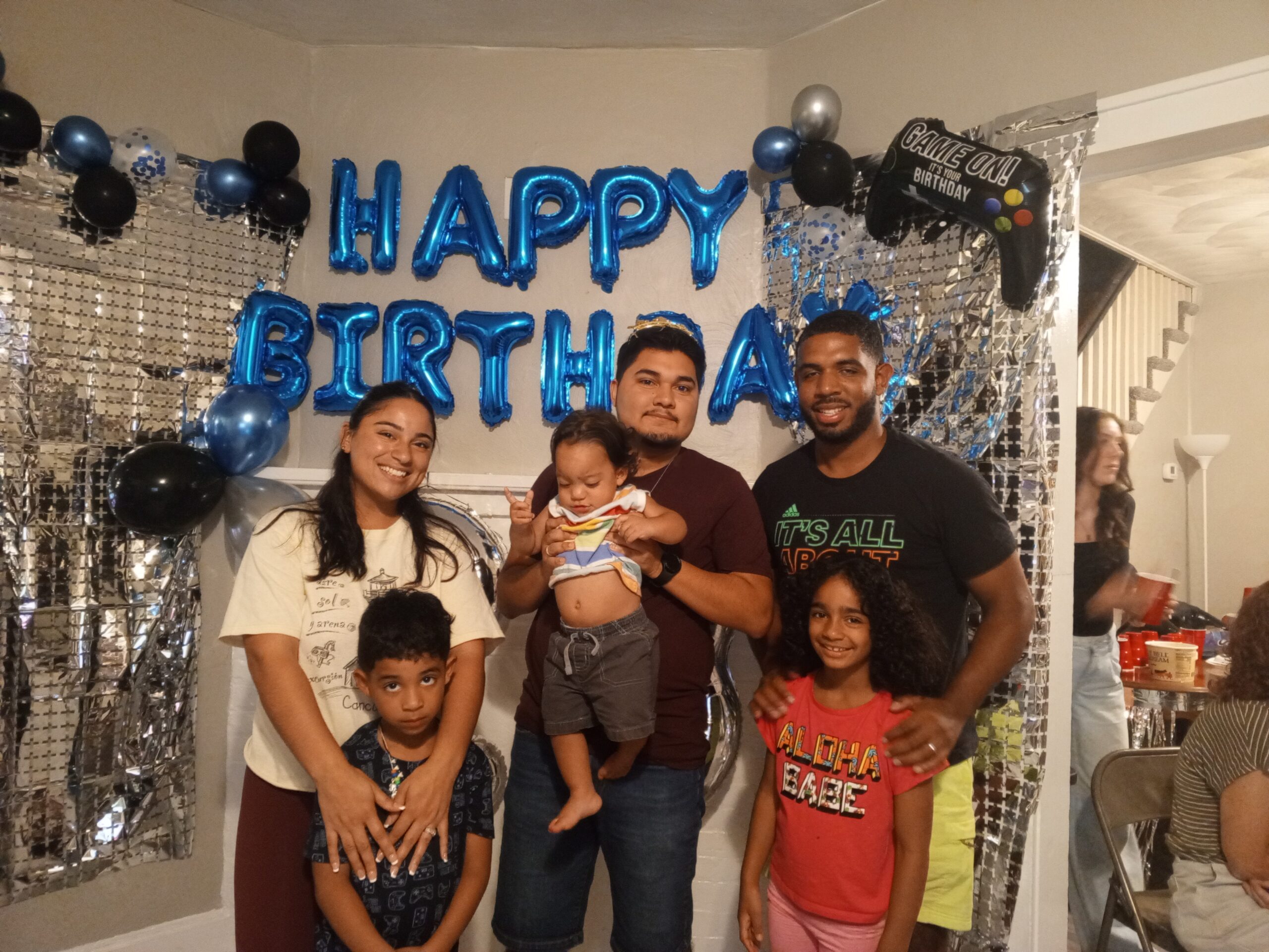 Family celebrating a birthday with "HAPPY BIRTHDAY" balloons, children and adults smiling, festive decorations, and a gaming-themed balloon in a lively indoor setting.