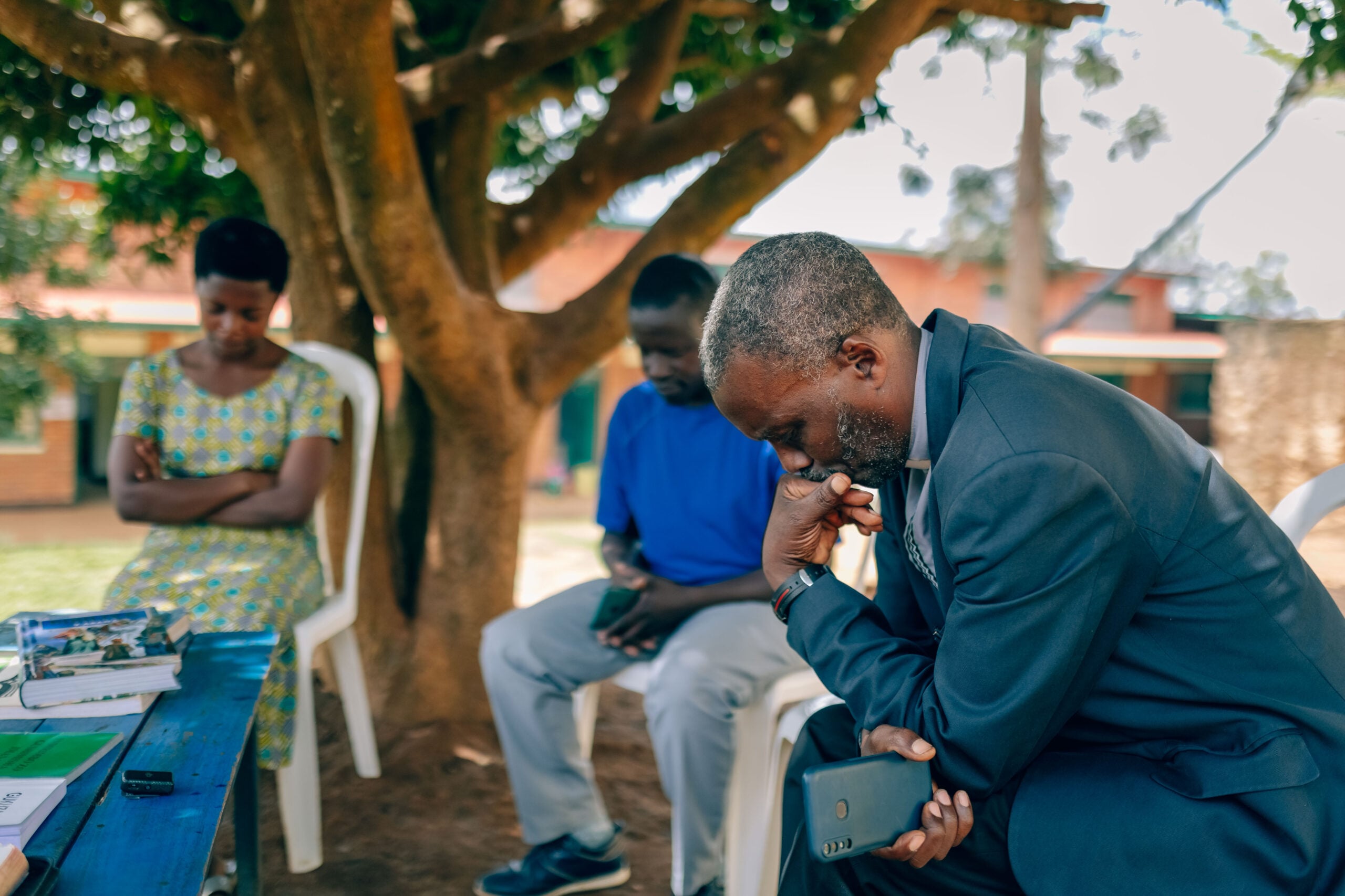 Pastor Jean deep in thought, seated under a tree with two others, reflecting on the challenges of church leadership and community support in Rwanda.