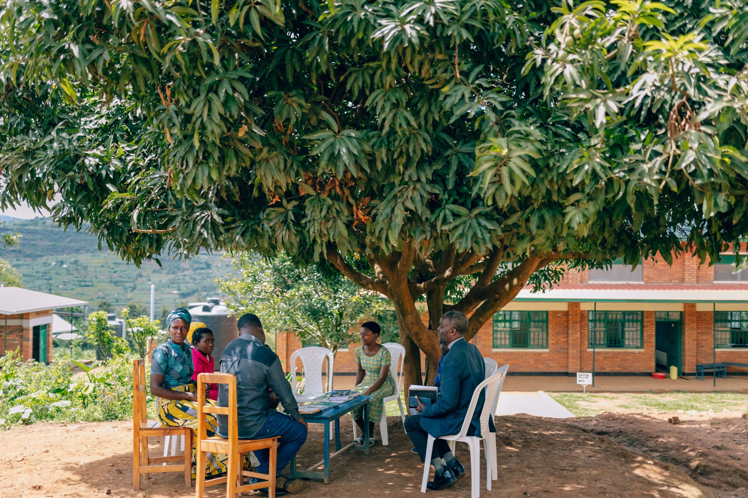 Group meeting under a large tree, discussing church multiplication and community support, with individuals in traditional attire and a building in the background, symbolizing resilience and revival efforts in rural Rwanda.