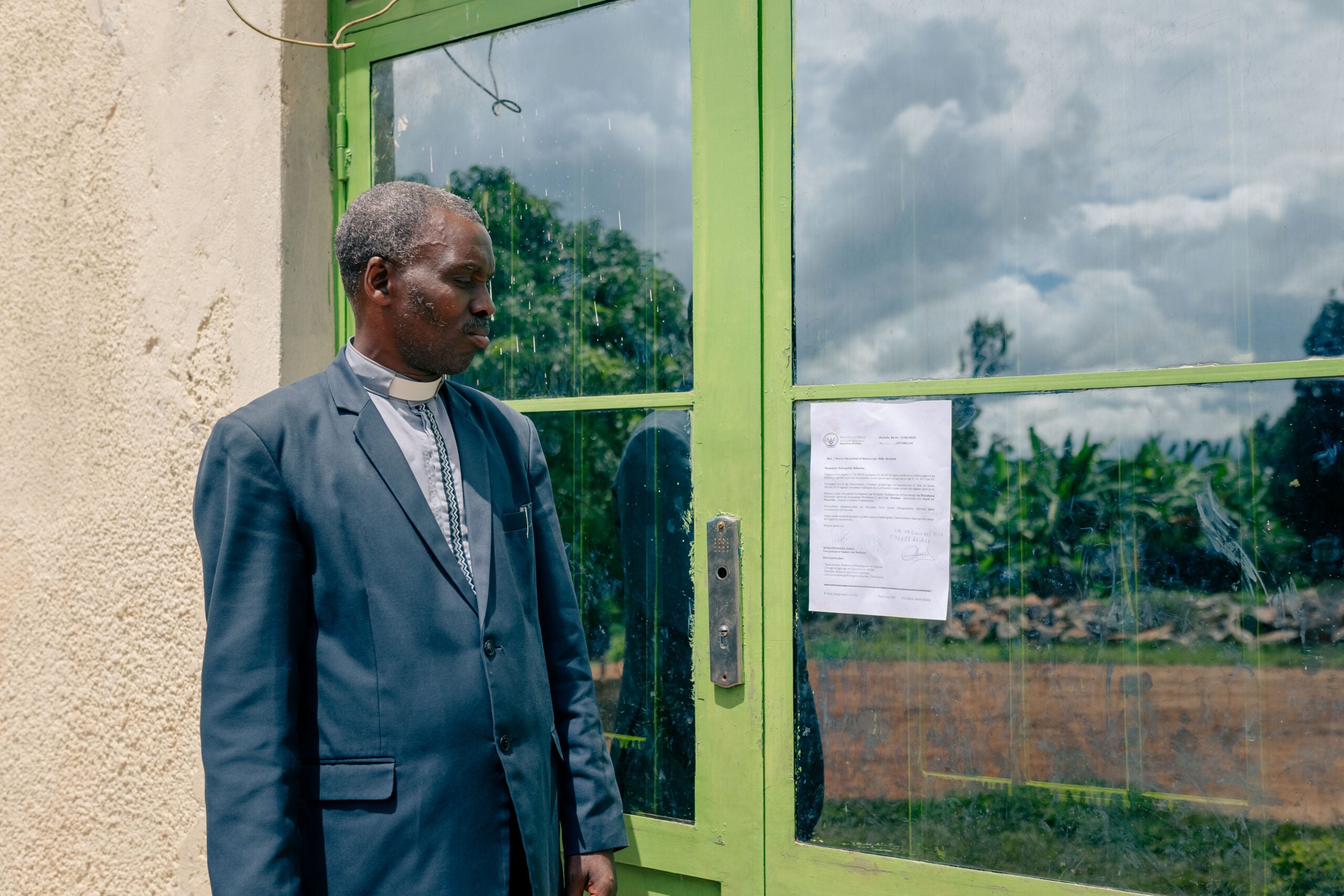 Pastor Jean standing in front of a green door with a notice, reflecting on the closure of his church building, symbolizing the challenges faced in leading a decentralized congregation.