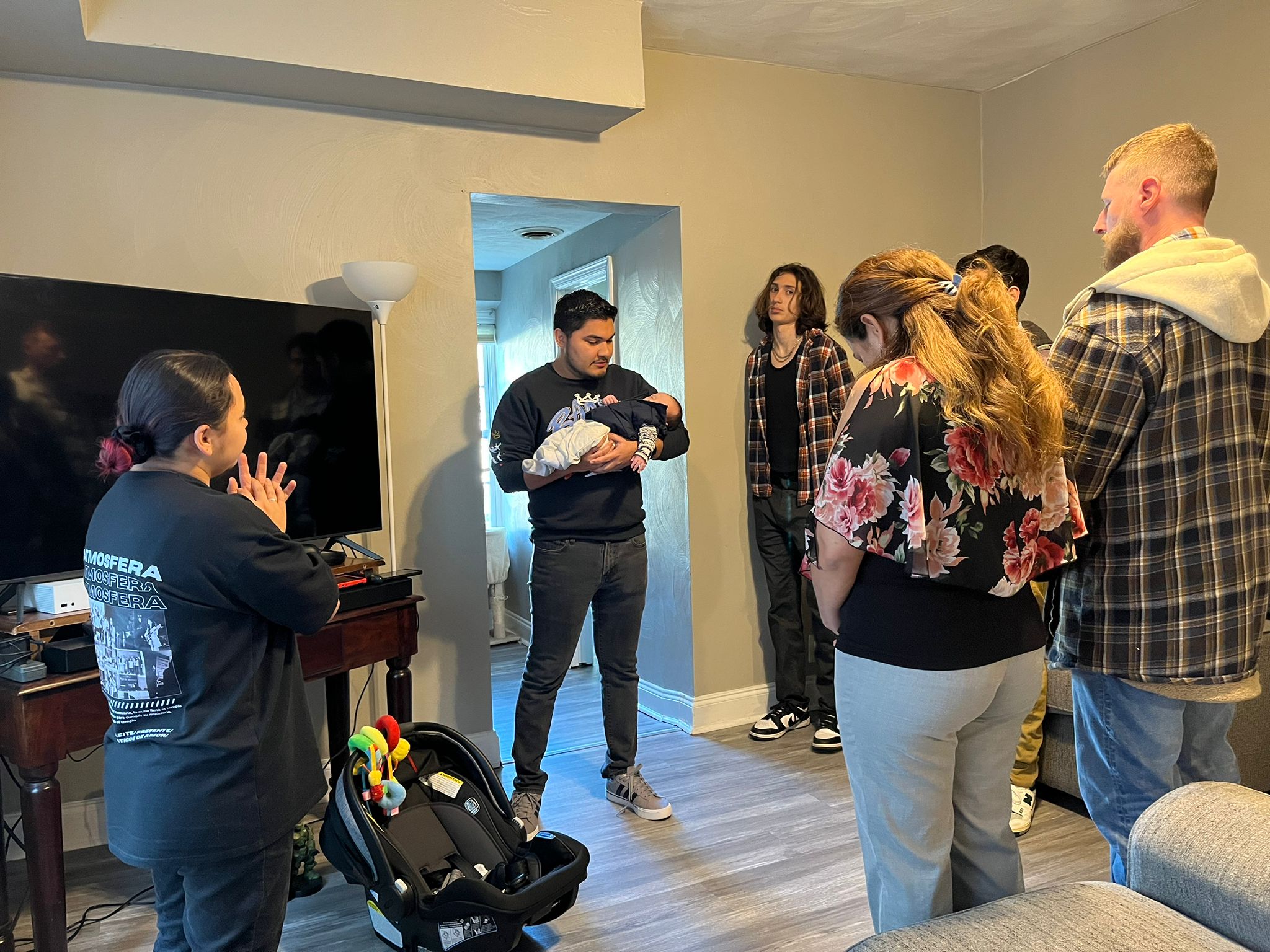 Group of people in a living room participating in a prayer gathering, with a man holding a baby, while others stand in a circle, demonstrating community support and connection among Spanish-speaking believers.