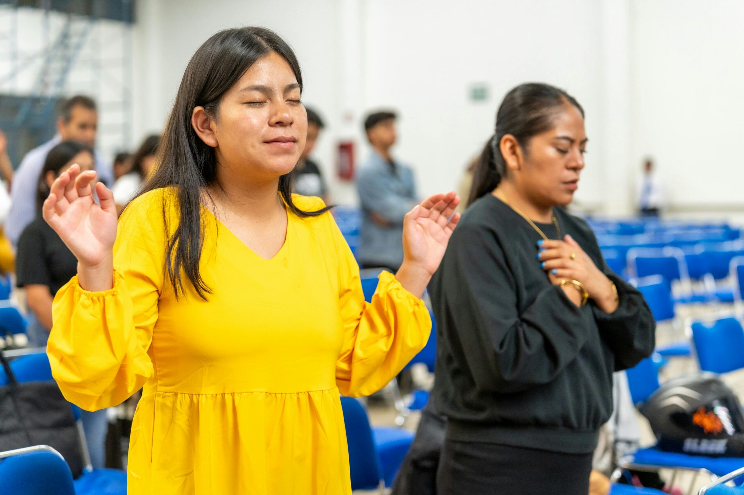 Women in prayer during a church service, expressing faith and community engagement, with blue chairs in the background, reflecting the mission of Iglesia Bautista Emanuel in Cuautla.