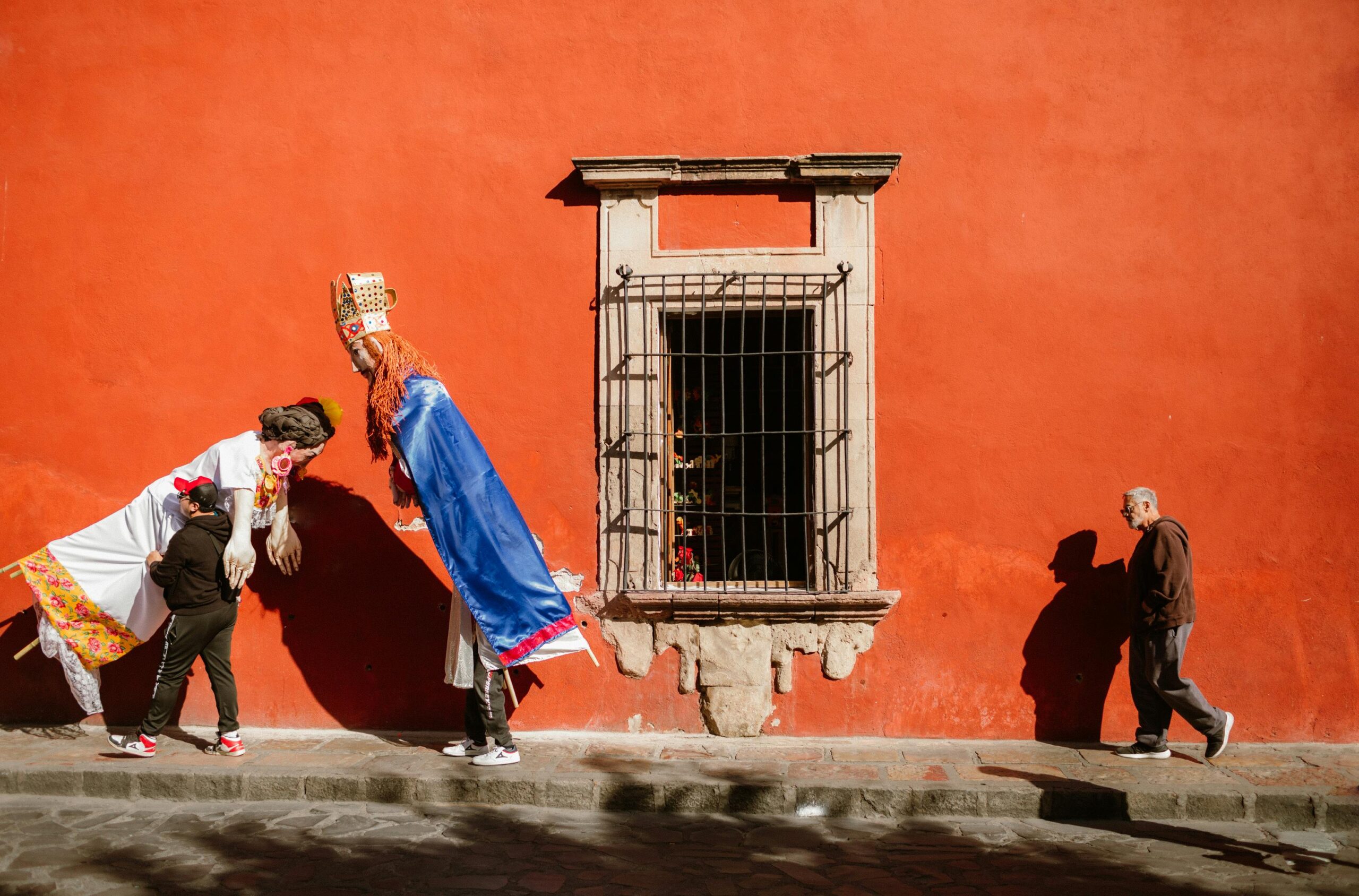 Two individuals dressed in large traditional costumes interacting on a vibrant orange wall, with an elderly man walking by, representing cultural celebration and community engagement in Cuautla, Mexico.