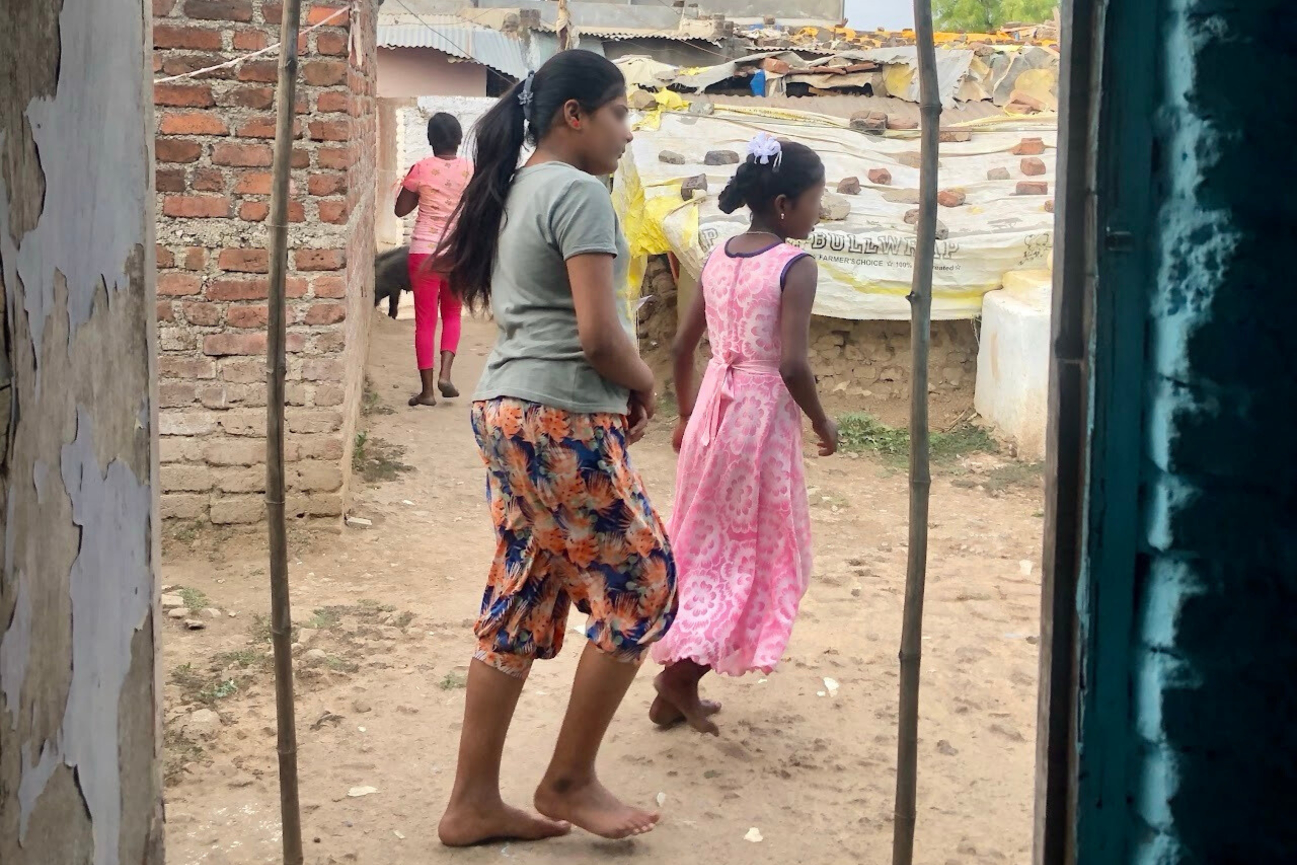 Two young girls walking barefoot in a rural setting, one in a gray shirt and patterned pants, the other in a pink dress, amidst a backdrop of modest homes and dirt paths, reflecting the community context of Pastor P.A.'s ministry in Amaravati.