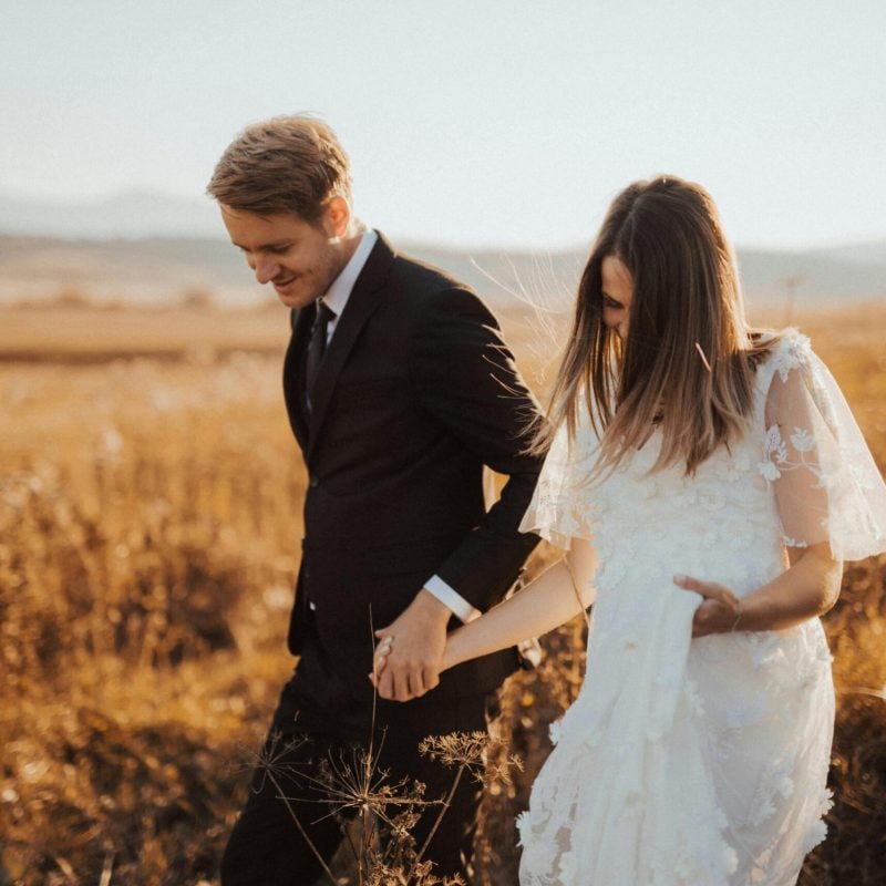 Couple walking hand in hand in a golden field, dressed in formal attire, symbolizing unity and partnership in a serene outdoor setting.